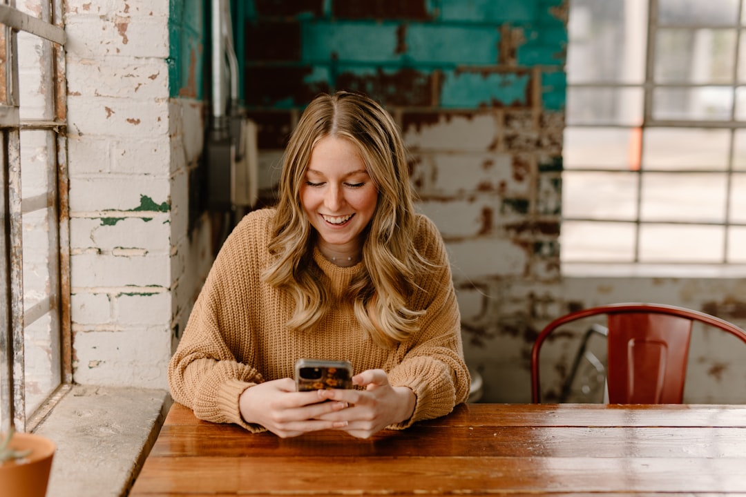 A woman sits at her kitchen table scrolling through photos on her phone, trying to pick one for a personalized gift for Mom